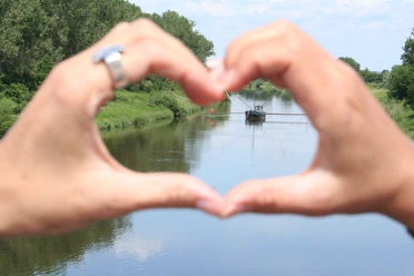 Hands form hearts and frame a traditional eel barge on a calm river.