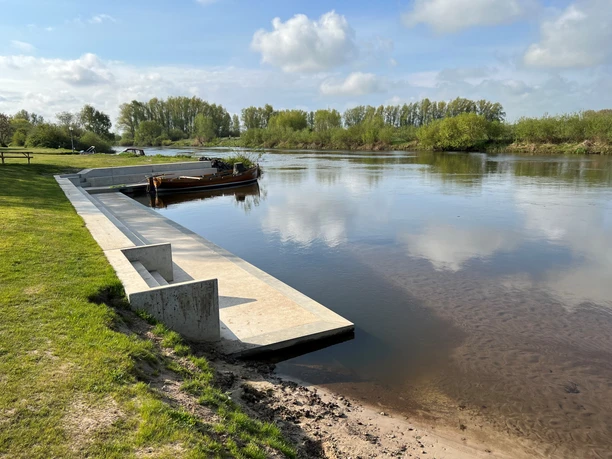 Bootsanleger aus Beton führt zum ruhigen Flussufer mit grüner Landschaft und bewölktem Himmel im Hintergrund.