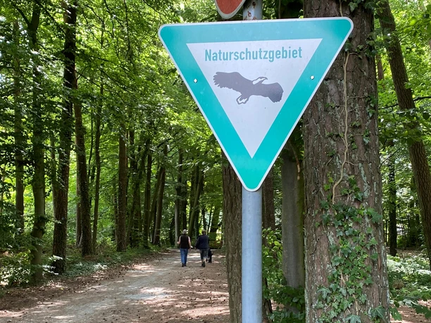 Naturschutzgebiet Foddenbach-Landbach Ein grünes Schild mit der Aufschrift "Naturschutzgebiet" steht an einem Waldweg, Personen gehen spazieren.