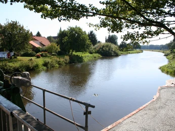Große Aue Liebenau Fluss mit sanften Ufern, umgeben von grünen Bäumen und einer kleinen Ortschaft im Hintergrund.