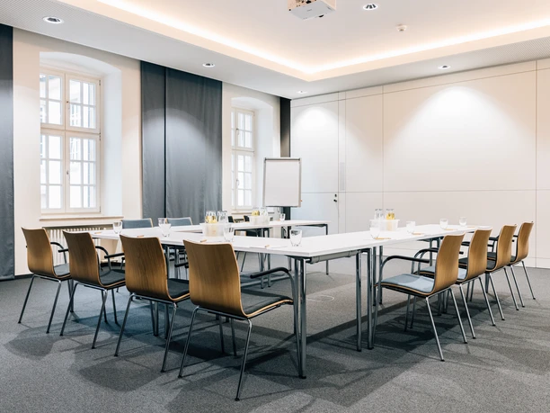 Moderner Tagungsraum mit hellem Tisch und zwölf Stühlen, ausgestattet mit Präsentationstechnik.