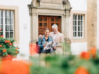 Ein Paar steht lächelnd vor einem historischen weißen Gebäude mit hölzerner Tür und Säulenelementen.