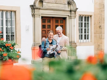 Ein Paar steht lächelnd vor einem historischen weißen Gebäude mit hölzerner Tür und Säulenelementen.