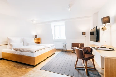Helles, modernes Hotelzimmer mit Doppelbett, Schreibtisch, Stuhl und Wandfernseher in stilvollem Design.