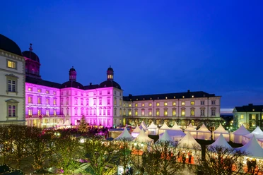 Schloß Bensberg Unikat-Weihnachtsmarkt Prächtig beleuchtetes Schloss mit violetten Lichtern bei Nacht, umgeben von stimmungsvollen Marktständen.