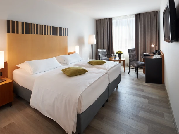 Doppelzimmer mit zwei weißen Betten, dunklen Vorhängen und einem Schreibtisch unter einem Fenster.