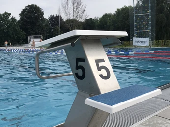 Springturm mit blauer Trittabdeckung am Rande eines Freibadbeckens, dahinter Bäume und Kletterwand.