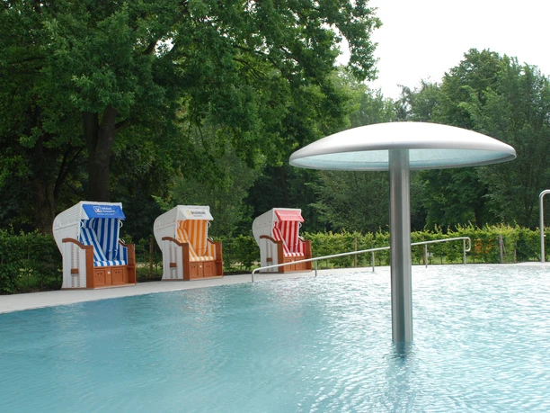 Ein ruhiges Schwimmbad mit einem Wassersprühpilz und bunten Strandkörben vor grüner Kulisse.