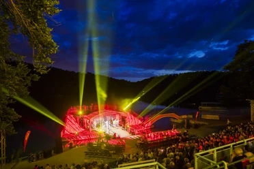 Seebühne Kriebstein - Kultur in Leipzig und Region Der Blick von den oberen Zuschauerrängen auf die Seebühne Kriebstein, ein Konzert wird gespielt, die Zuschauerplätze sind gefüllt und die Scheinwerfer strahlen in die verschiedenen Himmelsrichtungen zur blauen Stunde, Kultur, Veranstaltung