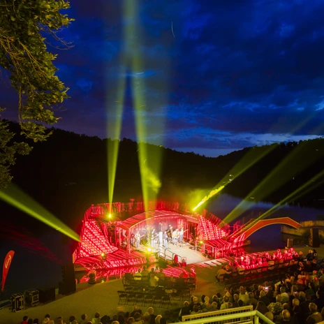 Widok na scenę nad jeziorem Kriebstein z górnych poziomów widowni, trwa koncert, miejsca są pełne, a reflektory świecą w różnych kierunkach o niebieskiej godzinie, kultura, wydarzenie