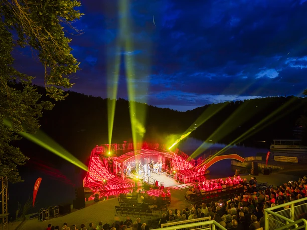 Widok na scenę nad jeziorem Kriebstein z górnych poziomów widowni, trwa koncert, miejsca są pełne, a reflektory świecą w różnych kierunkach o niebieskiej godzinie, kultura, wydarzenie