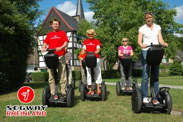 SEGWAYPoint-Haan-Rheinland Vier Personen fahren fröhlich auf Segways entlang eines grünen Wegs vor einem Fachwerkhaus.
