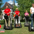 SEGWAYPoint-Haan-Rheinland Vier Personen fahren fröhlich auf Segways entlang eines grünen Wegs vor einem Fachwerkhaus.