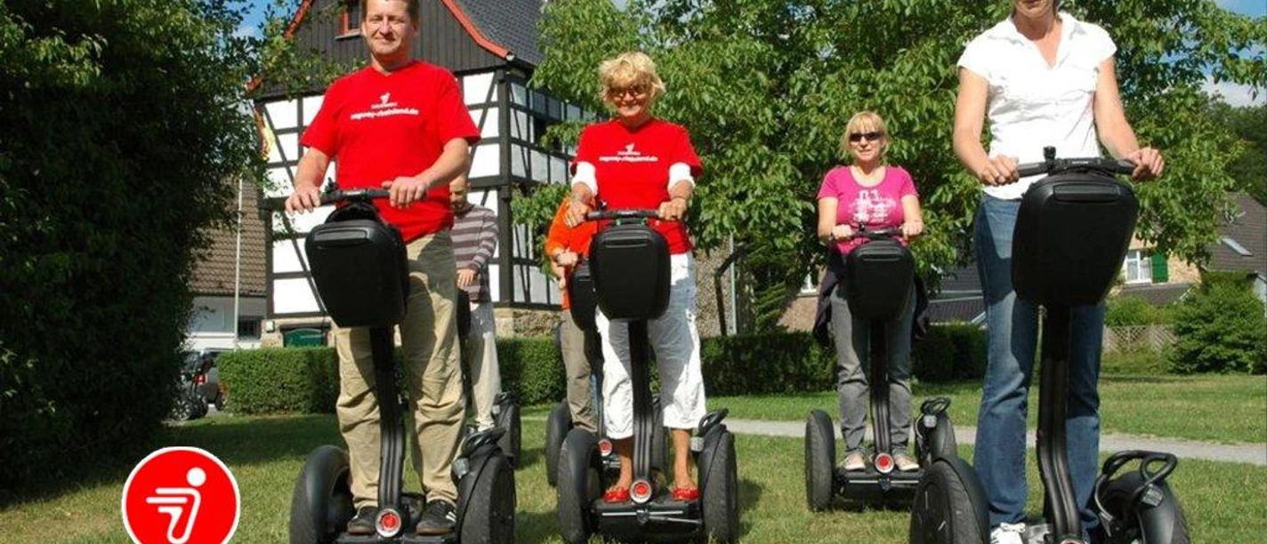 SEGWAYPoint-Haan-Rheinland Vier Personen fahren fröhlich auf Segways entlang eines grünen Wegs vor einem Fachwerkhaus.