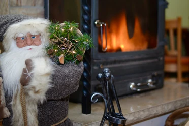 Een kerstfiguur met bril en grijze jas houdt een kleine dennentak vast naast een brandende open haard; een gezellige sfeer.