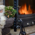 Kamin im Gasthaus Een kerstfiguur met bril en grijze jas houdt een kleine dennentak vast naast een brandende open haard; een gezellige sfeer.