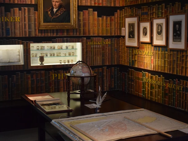 Das Bild zeigt einen Raum im Kultur- und Stadthistorischen Museum Duisburg. Die Wände sind mit detaillierten Illustrationen von Bücherregalen dekoriert, hinter denen themenspezifische Exponate beleuchtet ausgestellt sind. Vorne im Raum steht ein antiker Globus auf einem Tisch, umgeben von historischen Karten und einem Federkiel, was das historische Ambiente unterstreicht. An den Wänden sind Tafeln mit den Aufschriften „Heilkunde“, „Anatomie“ und „Römisches Recht“ zu sehen, die den thematischen Inhalt der Ausstellung anzeigen.