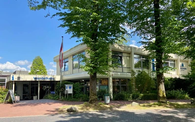 tourist-information-bad-zwischenahn-aussen.jpg Tourist-Information Bad Zwischenahn mit barrierefreiem Zugang und Beschilderung, umgeben von grünen Bäumen.