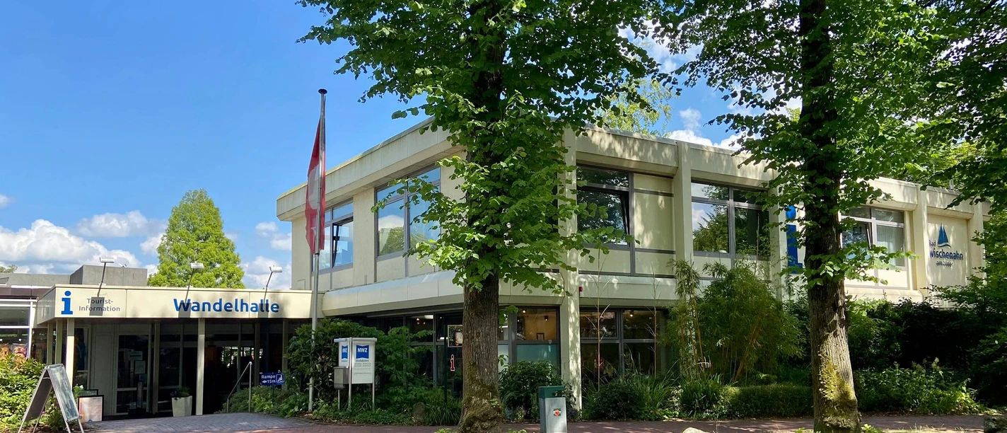 tourist-information-bad-zwischenahn-aussen.jpg Tourist-Information Bad Zwischenahn mit barrierefreiem Zugang und Beschilderung, umgeben von grünen Bäumen.