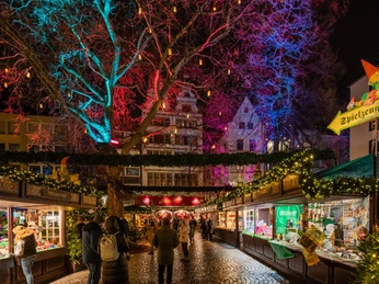 Heinzels Winter Fairytale Eine festlich erleuchtete Szenerie eines Weihnachtsmarktes bei Nacht. Bunte Lichter beleuchten die kahlen Äste eines großen Baumes, während dekorierte Marktstände Geschenke und Handwerkskunst präsentieren. Menschen bummeln entspannt über das Kopfsteinpflaster.A festively illuminated Christmas market scene at night. Colorful lights illuminate the bare branches of a large tree, while decorated market stalls display gifts and handicrafts. People stroll relaxed along the cobblestones.Een feestelijk verlicht kerstmarkttafereel bij nacht. Kleurrijke lichtjes verlichten de kale takken van een grote boom, terwijl versierde marktkraampjes geschenken en handwerk tentoonstellen. Mensen wandelen ontspannen over de kasseien.