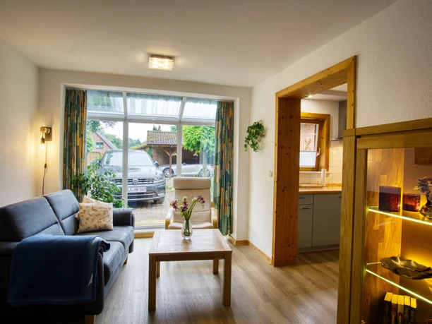 Wohnbereich.jpg Wohnbereich mit Sofa, Holztisch, großer Fensterfront und offenem Durchgang zur Küche.