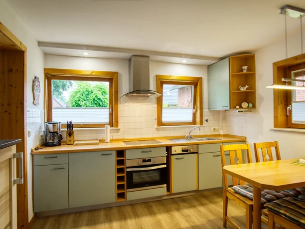 Küche.jpg Einbauküche mit Backofen, grünen Schränken und einem Holztisch mit Sitzkissen am Fenster.
