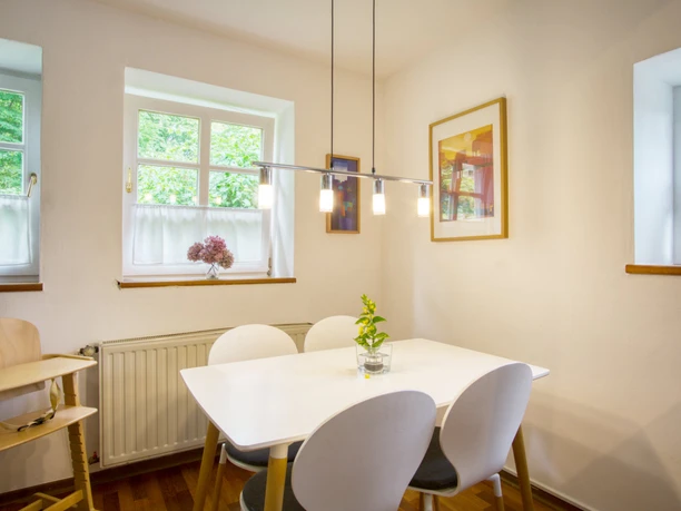 Essbereich.jpeg Heller Essplatz mit weißem Tisch, vier modernen Stühlen und einem Kinderhochstuhl aus Holz.