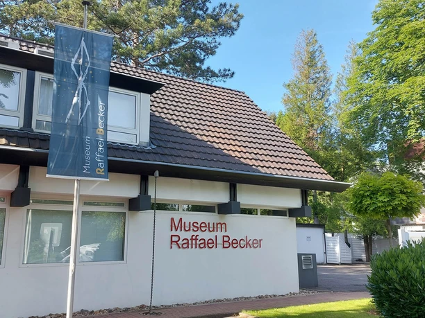Museum Raffael Becker The picture shows the bright façade of the modern Raffael Becker Museum. In front is a parking lot with a car. Rectangular windows and clear lines characterize the architecture, while the surrounding dense greenery breathes life into the building.