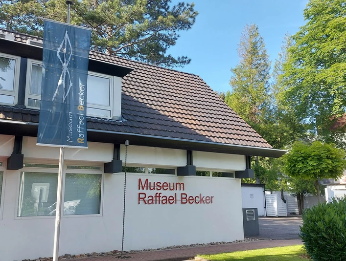 Museum Raffael Becker Das Bild zeigt die helle Fassade des modernen Museums Raffael Becker. Vorne ein Parkplatz mit Auto. Rechteckige Fenster und klare Linien prägen die Architektur, während umgebender, dichter Baumgrün dem Gebäude Leben einhaucht.The picture shows the bright façade of the modern Raffael Becker Museum. In front is a parking lot with a car. Rectangular windows and clear lines characterize the architecture, while the surrounding dense greenery breathes life into the building.