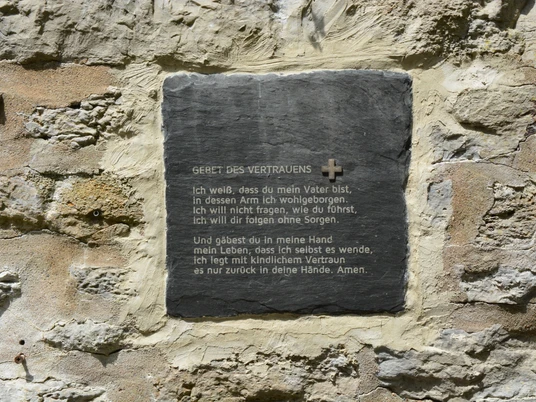 Eine Gedenktafel mit dem Gebet des Vertrauens ist in eine alte, verwitterte Steinwand eingebettet.