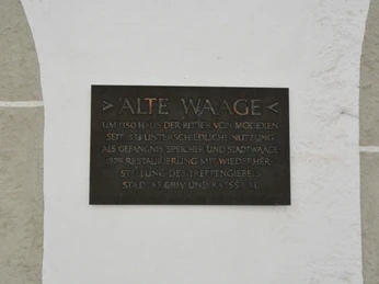 Metalltafel an einer hellen Wand, die die Geschichte der "Alten Waage" seit 1350 beschreibt.