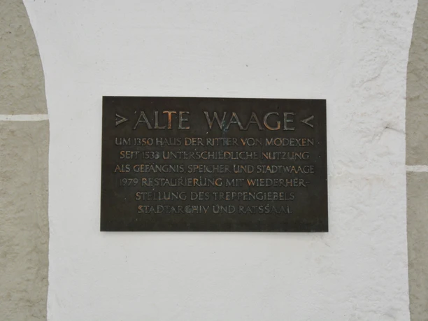 Metalltafel an einer hellen Wand, die die Geschichte der "Alten Waage" seit 1350 beschreibt.