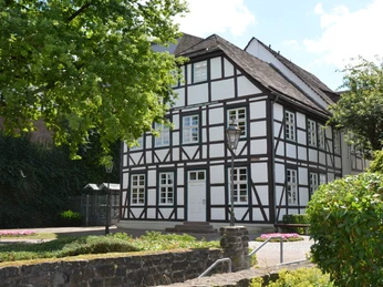 Haus des Gastes Fachwerkhaus mit weißer Fassade und dunklen Balken, umgeben von Grün, im Sonnenlicht liegend.