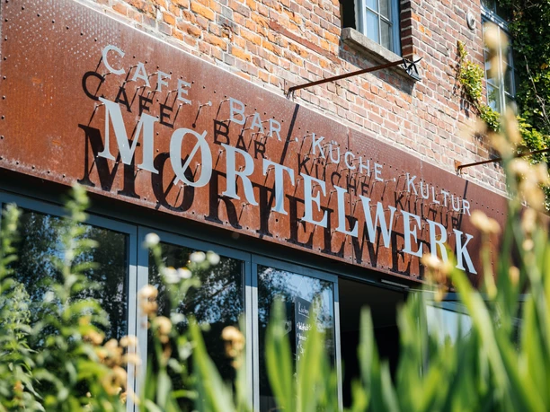 View of the brick façade with Moertelwerk lettering, surrounded by plants, gastronomy, outdoor seating