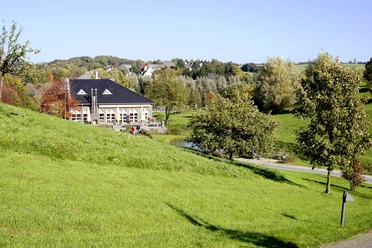 Golf Club Grevenmühle in Ratingen Ein modernes Clubhaus liegt eingebettet in eine weitläufige, grüne Parklandschaft mit vereinzelten Bäumen.