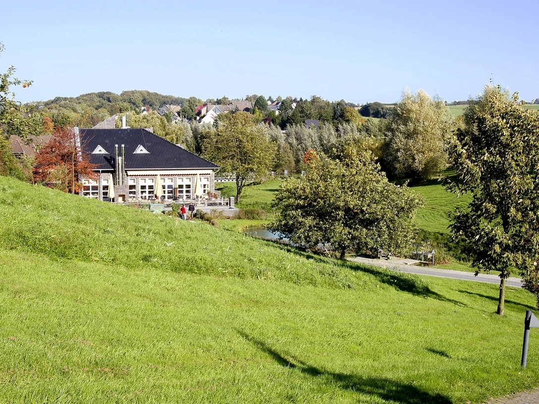 Golf Club Grevenmühle in Ratingen Ein modernes Clubhaus liegt eingebettet in eine weitläufige, grüne Parklandschaft mit vereinzelten Bäumen.