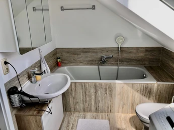 Gästezimmer Haus Beate Helles Badezimmer mit Dachschräge, freistehender Badewanne, Waschbecken und Spiegelwand.