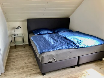 Gästezimmer Haus Beate Dachzimmer mit modernem Doppelbett, blauen Bettbezügen und Nachttischlampe auf Holzboden.