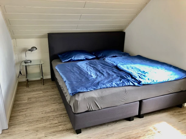 Gästezimmer Haus Beate Dachzimmer mit modernem Doppelbett, blauen Bettbezügen und Nachttischlampe auf Holzboden.