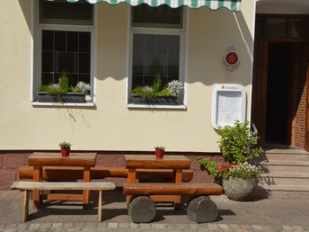 Zwei rustikale Holzbänke und Tische vor einem Gasthofeingang mit Blumenschmuck und Markise.