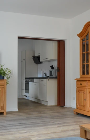 Wohnzimmer mit Holzmöbeln und Blick in eine moderne Küche; heller Holzfußboden, Pflanzen im Raum.