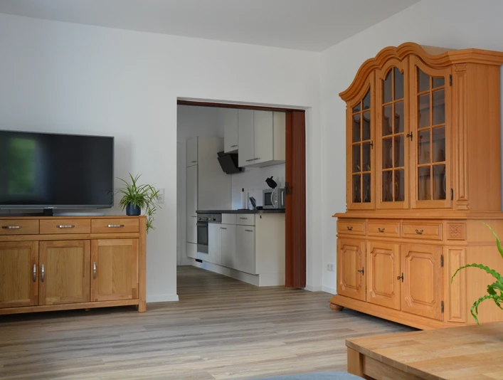 Wohnzimmer mit Holzmöbeln und Blick in eine moderne Küche; heller Holzfußboden, Pflanzen im Raum.