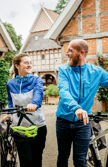 Radfahren auf der Artland-Rad-Tour Lächelndes Paar mit Fahrrädern vor historischen Fachwerkgebäuden in ländlicher Umgebung.Smiling couple with bicycles in front of historic half-timbered buildings in a rural setting.Smilende par med cykler foran historiske bindingsværksbygninger i landlige omgivelser.Lachend stel met fietsen voor historische vakwerkhuizen in een landelijke omgeving.