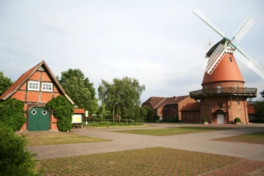 Rote Backsteinmühle mit weißen Flügeln neben grünem Fachwerkhaus, umgeben von Bäumen und Weg.