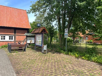 Mühlenplatz Landesbergen Ein rustikales rotes Backsteinhaus mit Infotafeln und Bänken umgeben von Bäumen bei Sonnenschein.