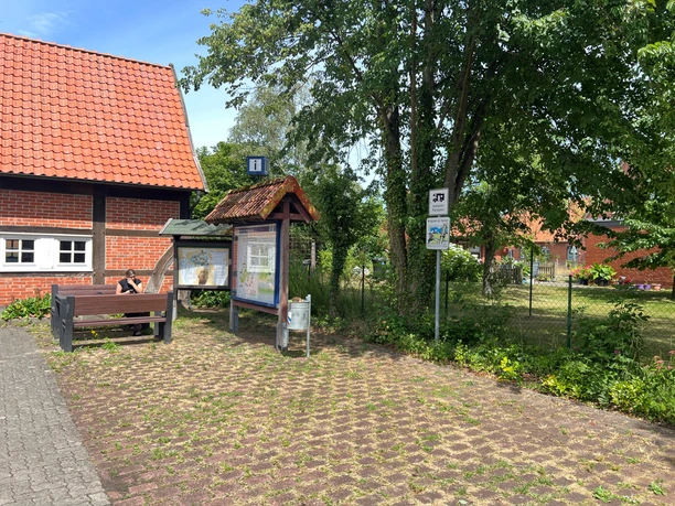 Mühlenplatz Landesbergen Ein rustikales rotes Backsteinhaus mit Infotafeln und Bänken umgeben von Bäumen bei Sonnenschein.