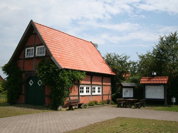 Backsteingebäude mit rotem Ziegeldach, umgeben von Grün, bei leicht bewölktem Himmel auf dem Mühlenplatz.