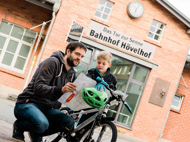 Mann und Kind betrachten eine Karte vor dem Backsteingebäude des Bahnhofs Hövelhof.