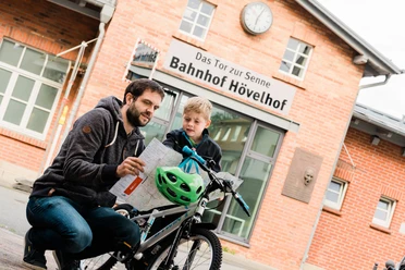 Bahnhof Hövelhof - Das Tor zur Senne Mann und Kind betrachten eine Karte vor dem Backsteingebäude des Bahnhofs Hövelhof.