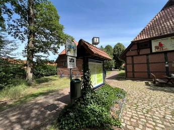 Wohnmobilstellplatz mit Informationsschild und Fachwerkgebäuden, umgeben von Bäumen und gepflasterten Wegen.